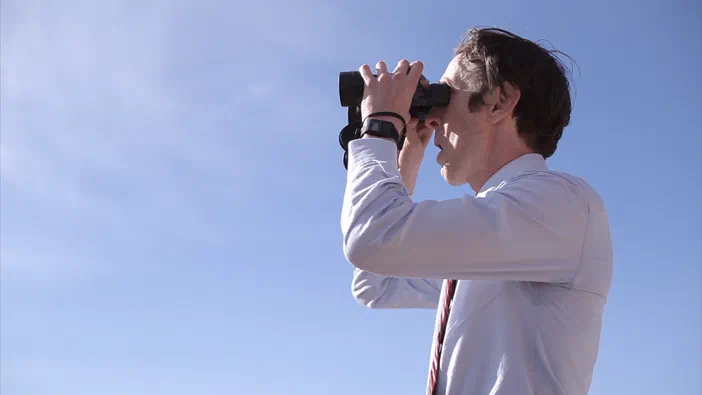 Un homme de profil portant une chemise blanche observe à travers des jumelles devant un ciel bleu clair.