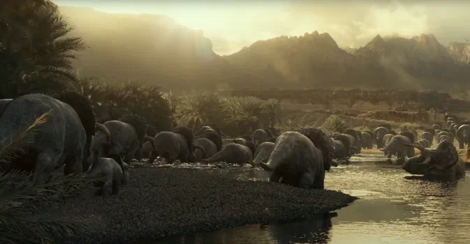 Un groupe de dinosaures herbivores à crête rouge se baigne dans une rivière au sein d'un paysage préhistorique montagneux.