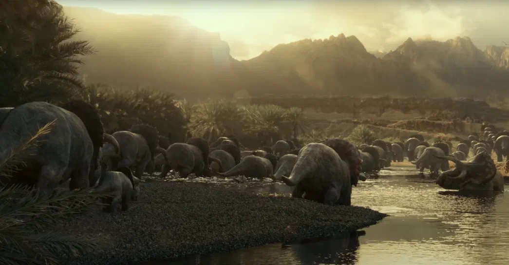 Un groupe de dinosaures herbivores à crête rouge se baigne dans une rivière au sein d'un paysage préhistorique montagneux.