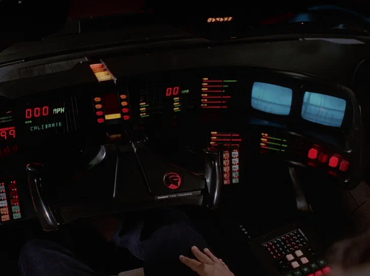 Vue intérieure du cockpit de la voiture K.I.T.T. avec son tableau de bord lumineux affichant des indicateurs rouges et verts, une vitesse à zéro et un logo distinctif au centre.