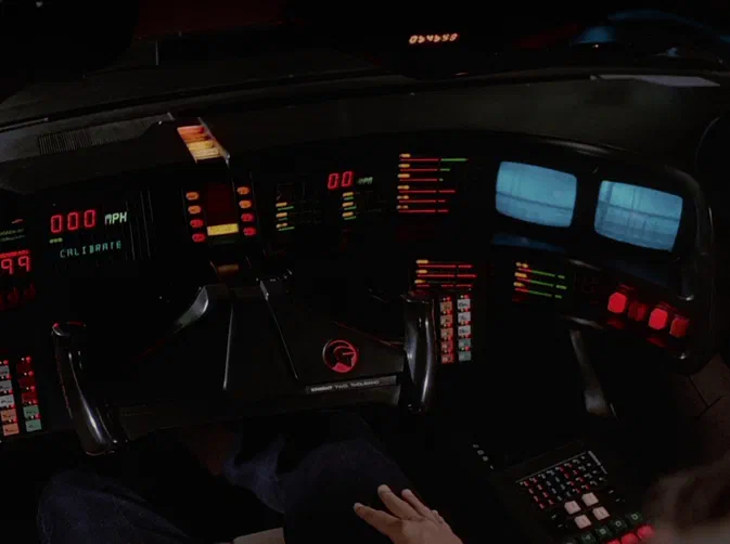 Vue intérieure du cockpit de la voiture K.I.T.T. avec son tableau de bord lumineux affichant des indicateurs rouges et verts, une vitesse à zéro et un logo distinctif au centre.