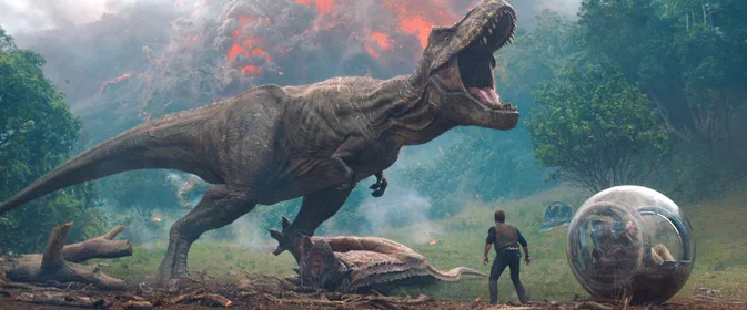 Dans Jurassic World : Le Royaume Chuté, un T-Rex rugit face à une éruption volcanique alors qu'un homme observe depuis près d'une sphère de verre.