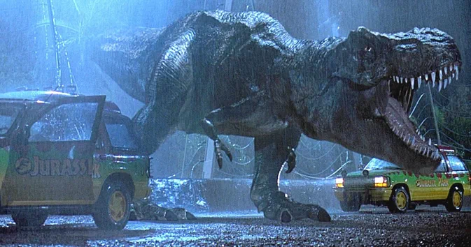 Un T-Rex géant se tient entre deux véhicules verts portant l'inscription 'Jurassic Park', sous la pluie dans une scène de film.