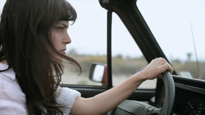 Une femme aux cheveux longs conduit une voiture dans un paysage désertique.